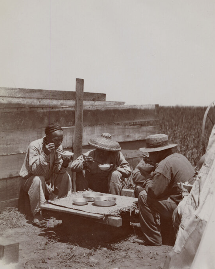 4 chinese farmworkers sit around a low outdoor table sharing a meal